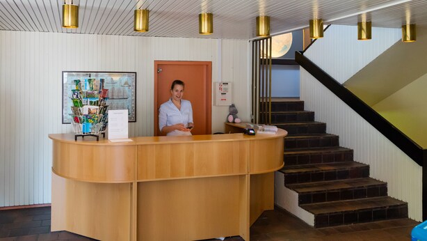 A reception desk with a wooden finish, a person standing behind it, and a staircase with a black railing leading to an upper level.