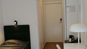 A small, neatly arranged room with a bed, a desk with a lamp, and a door.
