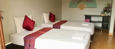 A hotel room with two beds, each with white linens and red pillows. There is a desk with a chair, a small shelf with a vase of flowers, and a wall-mounted fan.