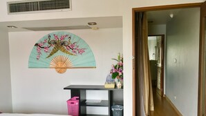 Une chambre avec un lit, un bureau sur lequel se trouve un panier à linge rose, un ventilateur fixé au mur et une décoration murale.