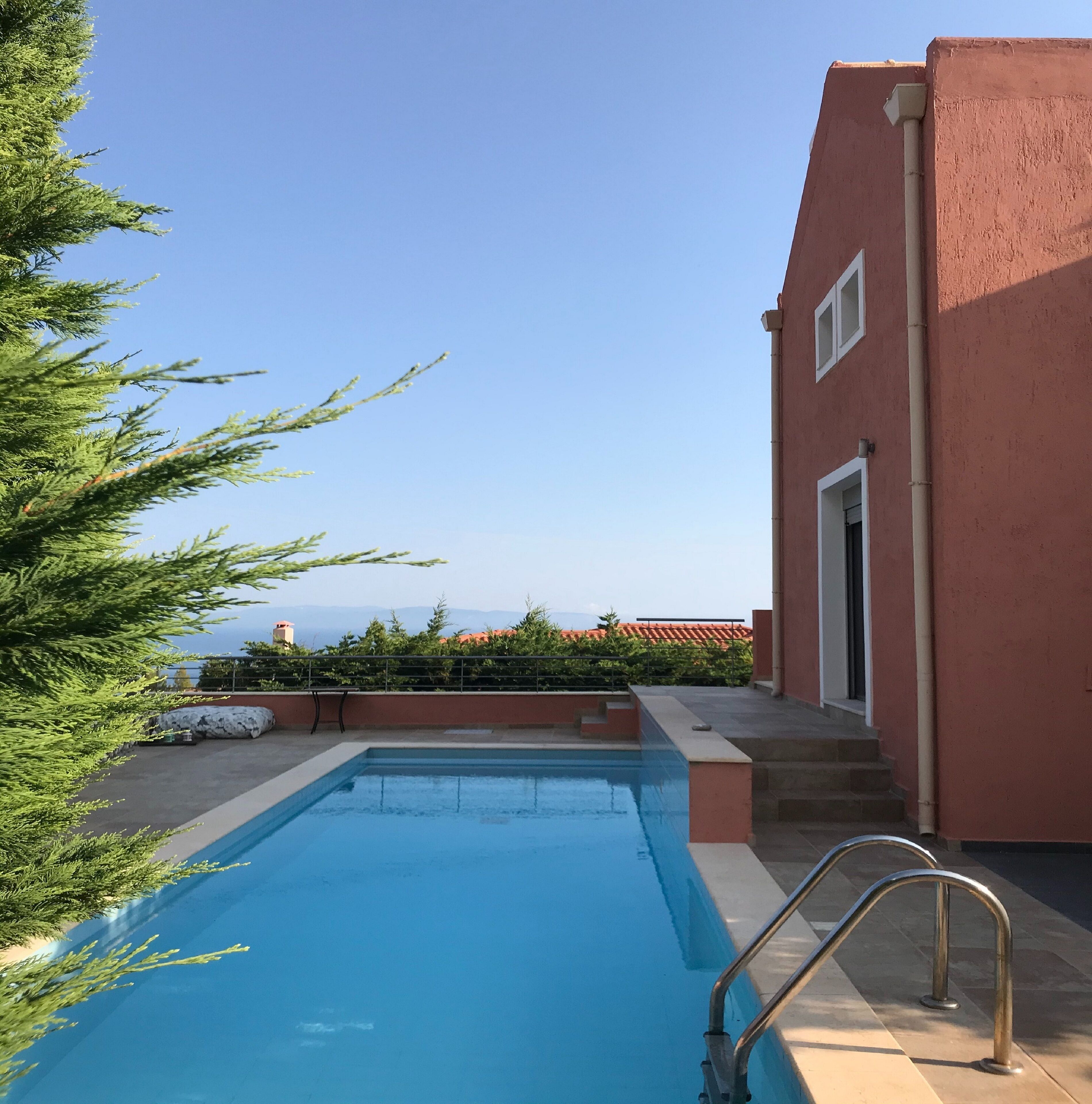 A swimming pool with a metal ladder, surrounded by a red building and greenery.