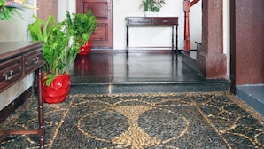 Un couloir avec un sol à motifs, une porte en bois, une console sur laquelle est posée une plante, et un vase rouge.