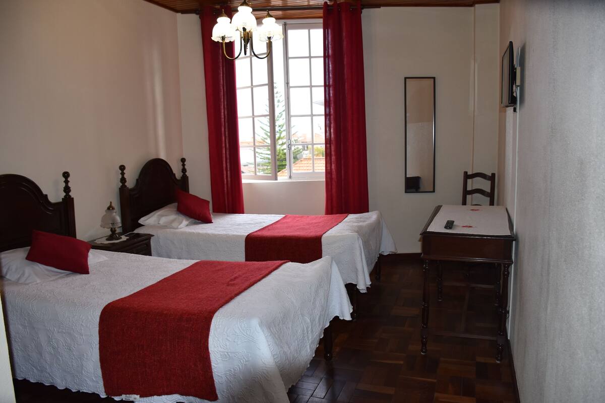 Une chambre d’hôtel avec deux lits, un plancher en bois, un bureau et une fenêtre avec des rideaux rouges.