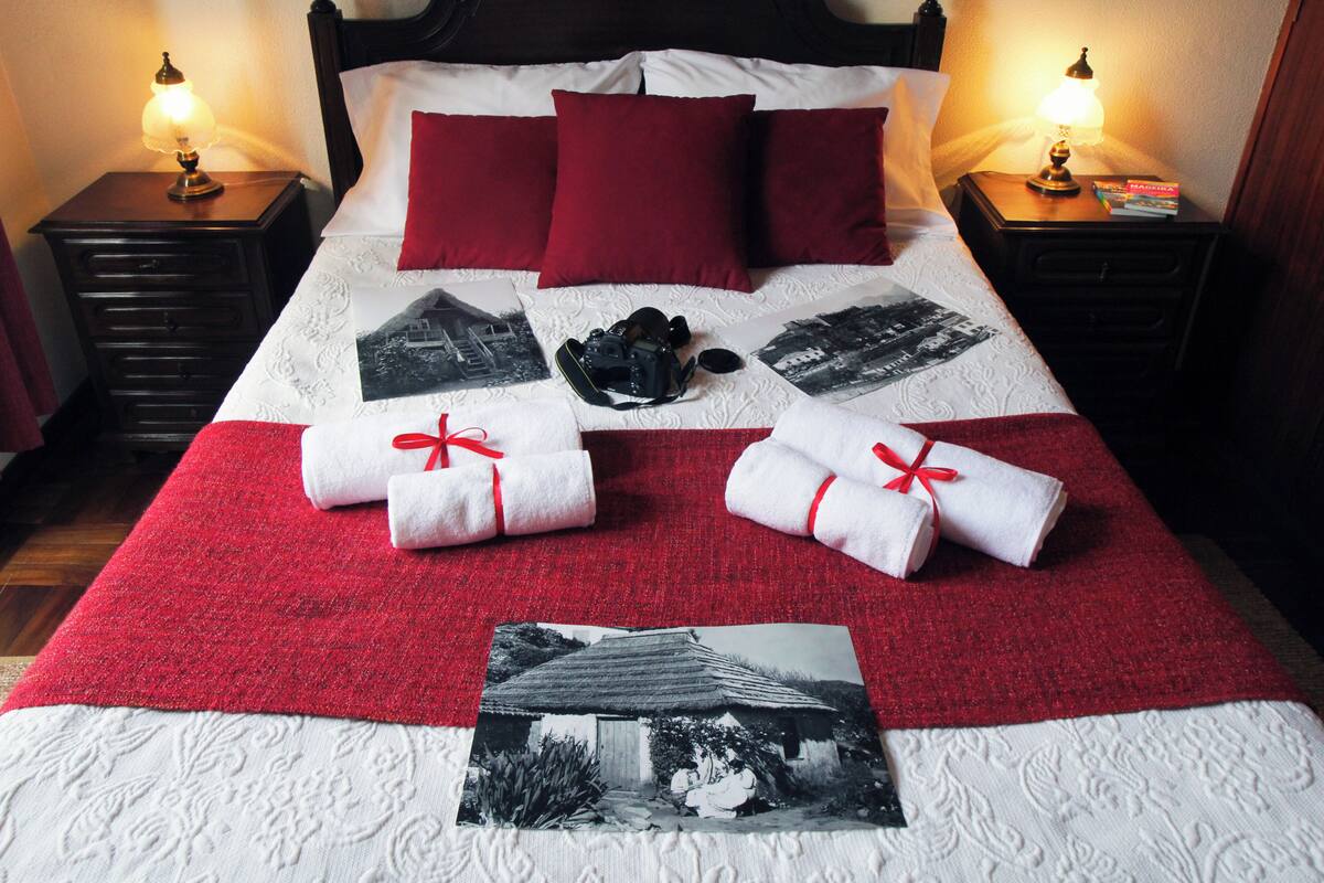 Un lit bien fait, recouvert d’une couverture rouge et blanche, avec des oreillers rouges et une photo d’une maison. Il y a deux tables de chevet, chacune avec une lampe, et un livre est posé sur l’une d’elles.