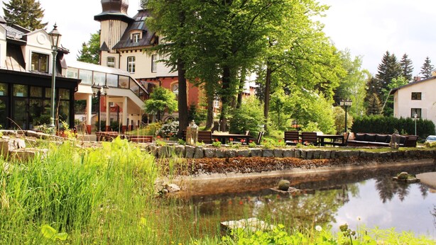 A serene riverside setting with a historic building featuring a tower, lush greenery, and a calm waterway. - Hotel Szrenicowy Dwór (Szklarska Poreba)