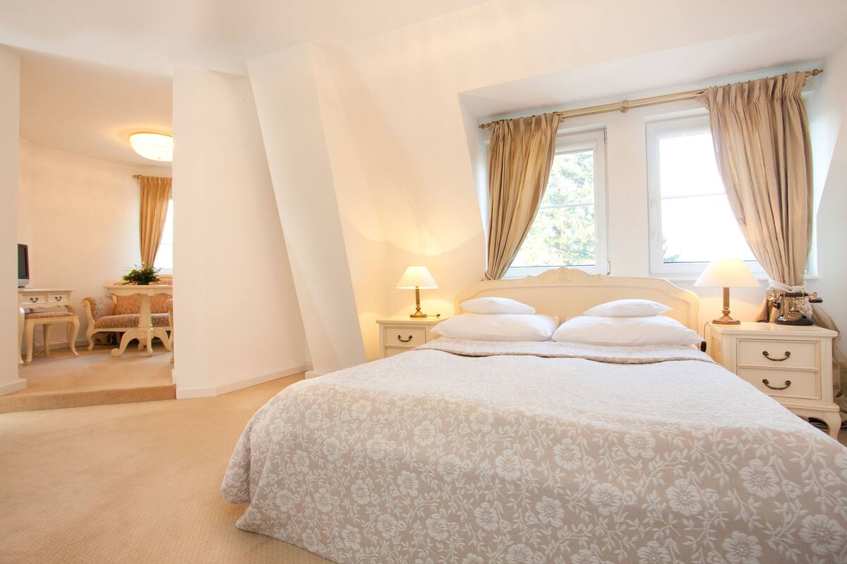 A bedroom with a large bed, two bedside tables with lamps, a desk with a chair, and a window with curtains.