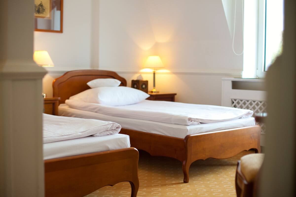 A hotel room with two wooden beds, each with white linens. There are bedside lamps and a small table with a lamp.