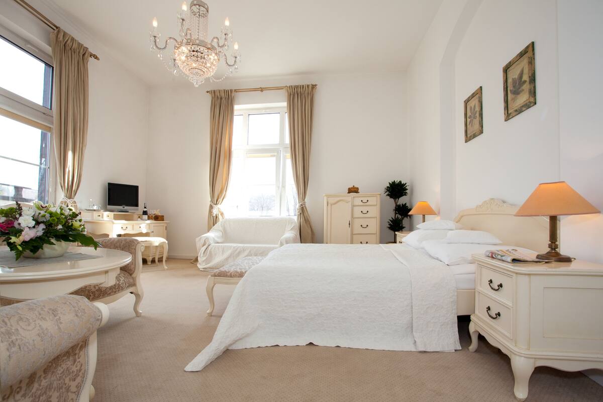 A hotel room with a large bed, a desk with a computer, a chair, a bedside table with a lamp, and a window with curtains.