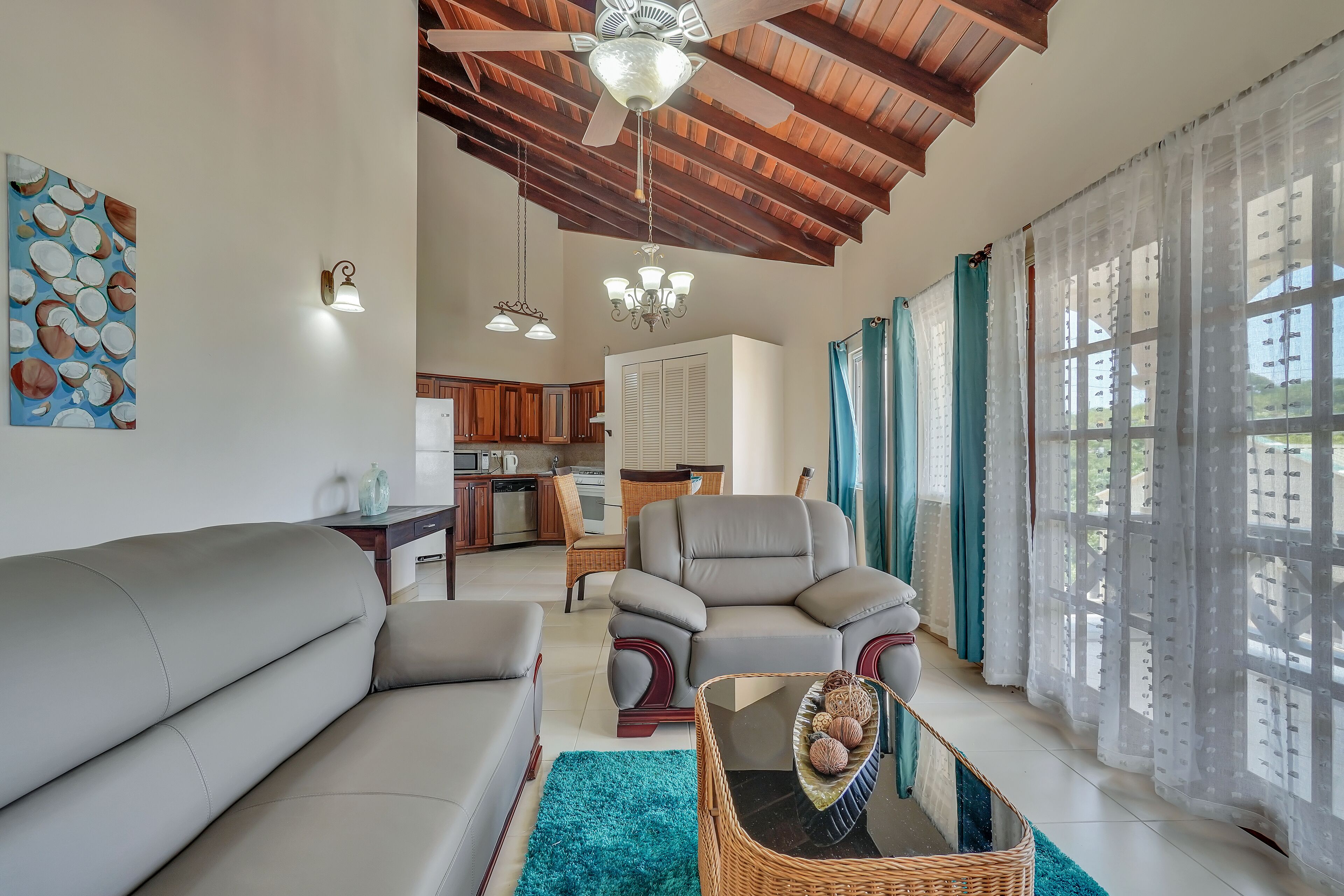 A spacious living room with a sofa, armchair, and a coffee table. There is a ceiling fan, a chandelier, and a view of a kitchen and dining area through large windows with sheer curtains.