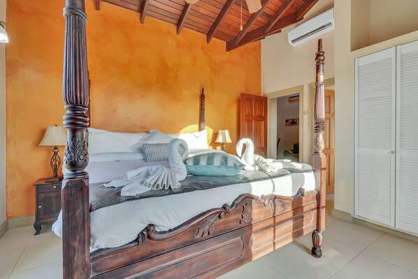 Una cama con dosel y cabecera de madera, una colcha blanca y almohadas decorativas. Hay una mesita de noche con una lámpara, un armario y una puerta que da a otra habitación.