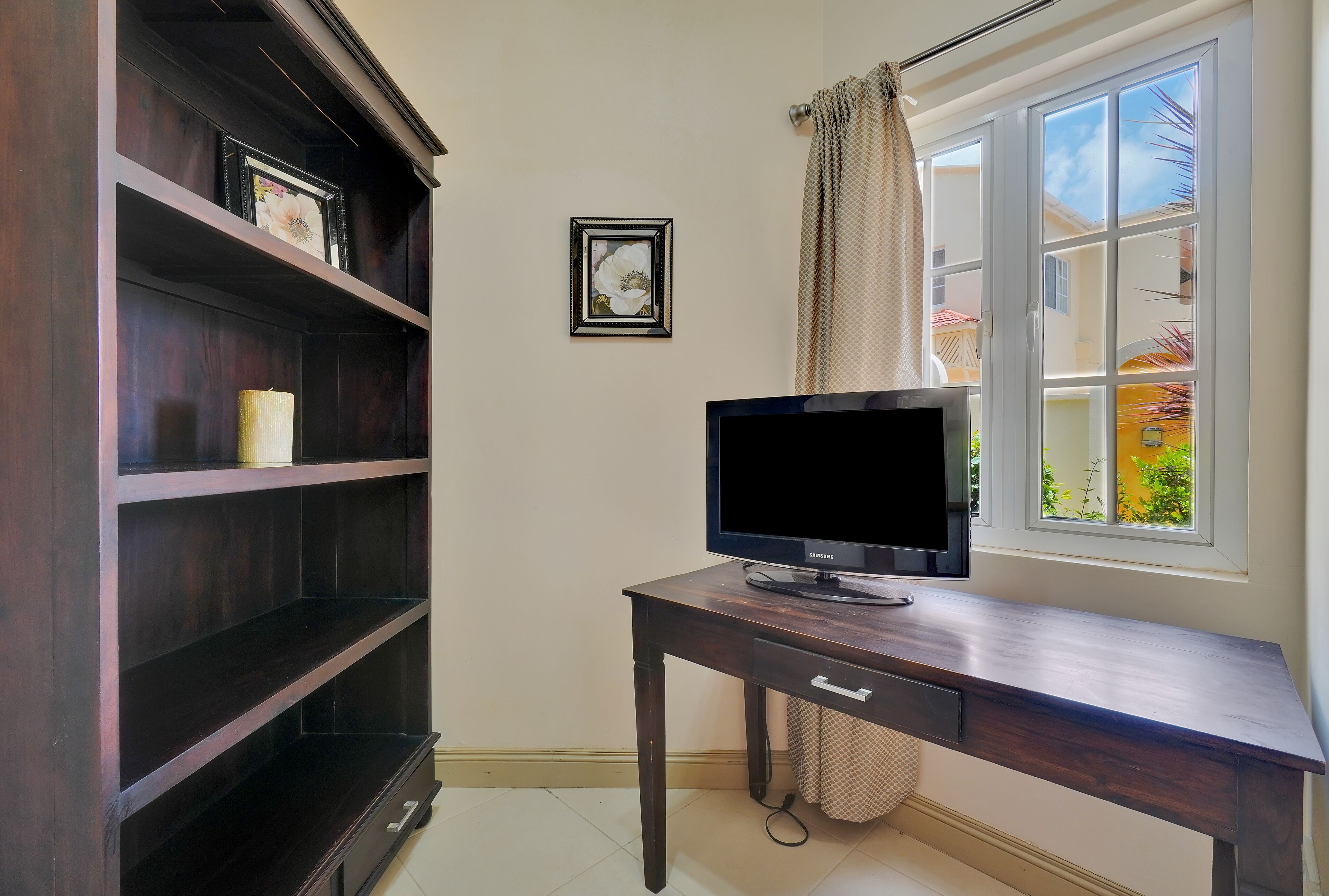 Un escritorio de madera oscura con un televisor de pantalla plana, una estantería con un cuadro enmarcado y una ventana con cortinas.