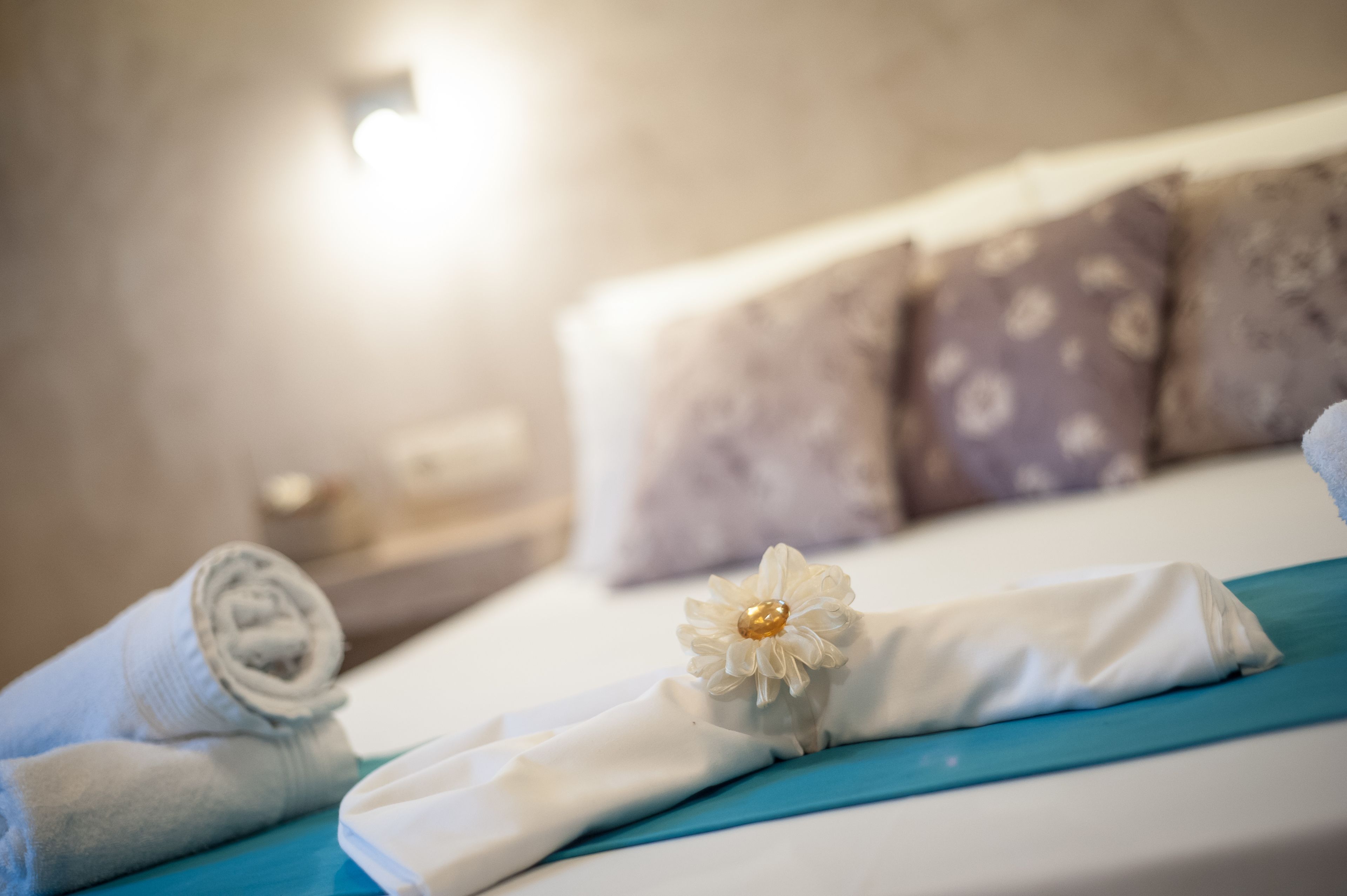 A neatly made bed with a blue bedspread, a rolled white towel, and a decorative flower.
