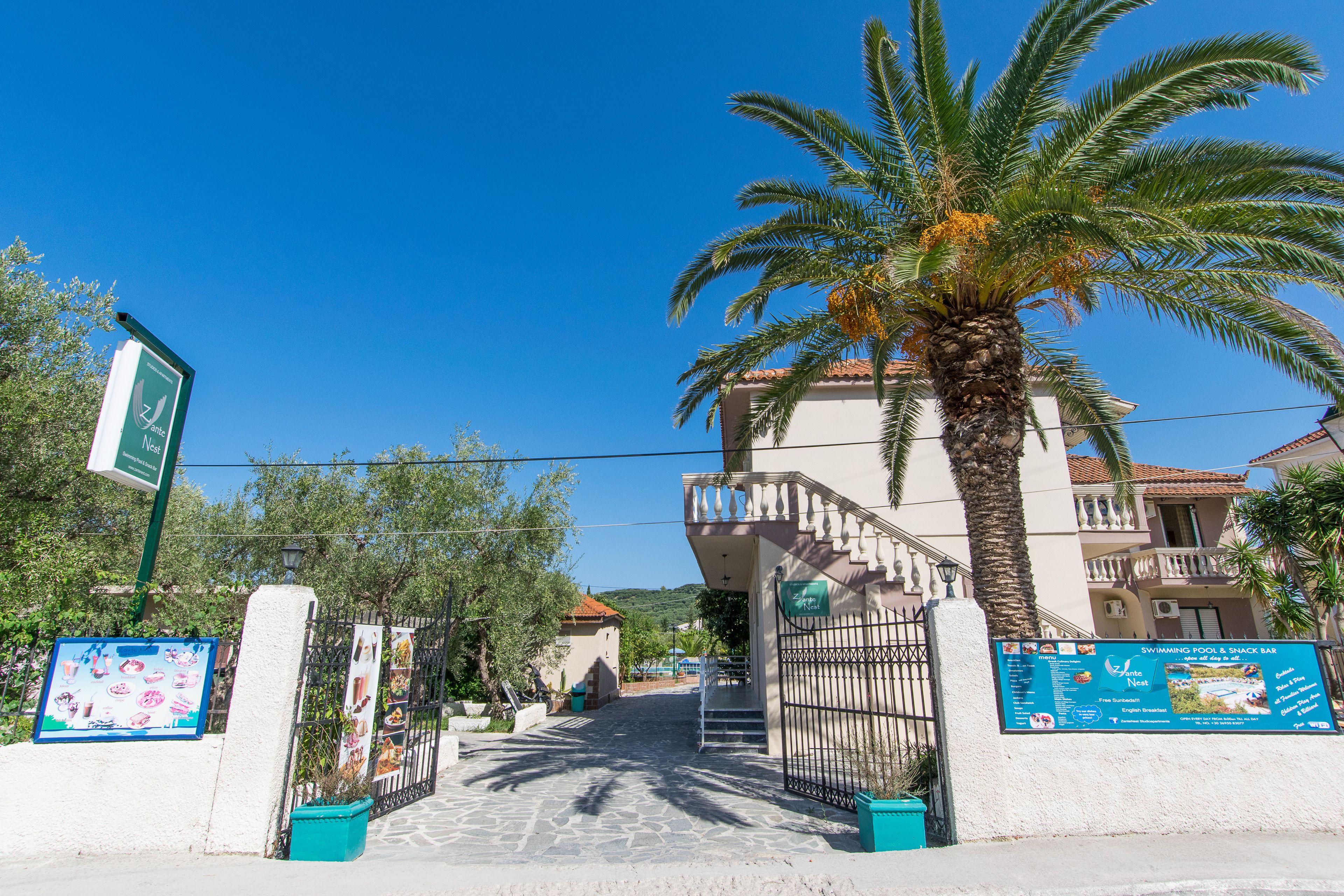 A gated entrance with a palm tree, a signboard, and a building with a balcony.