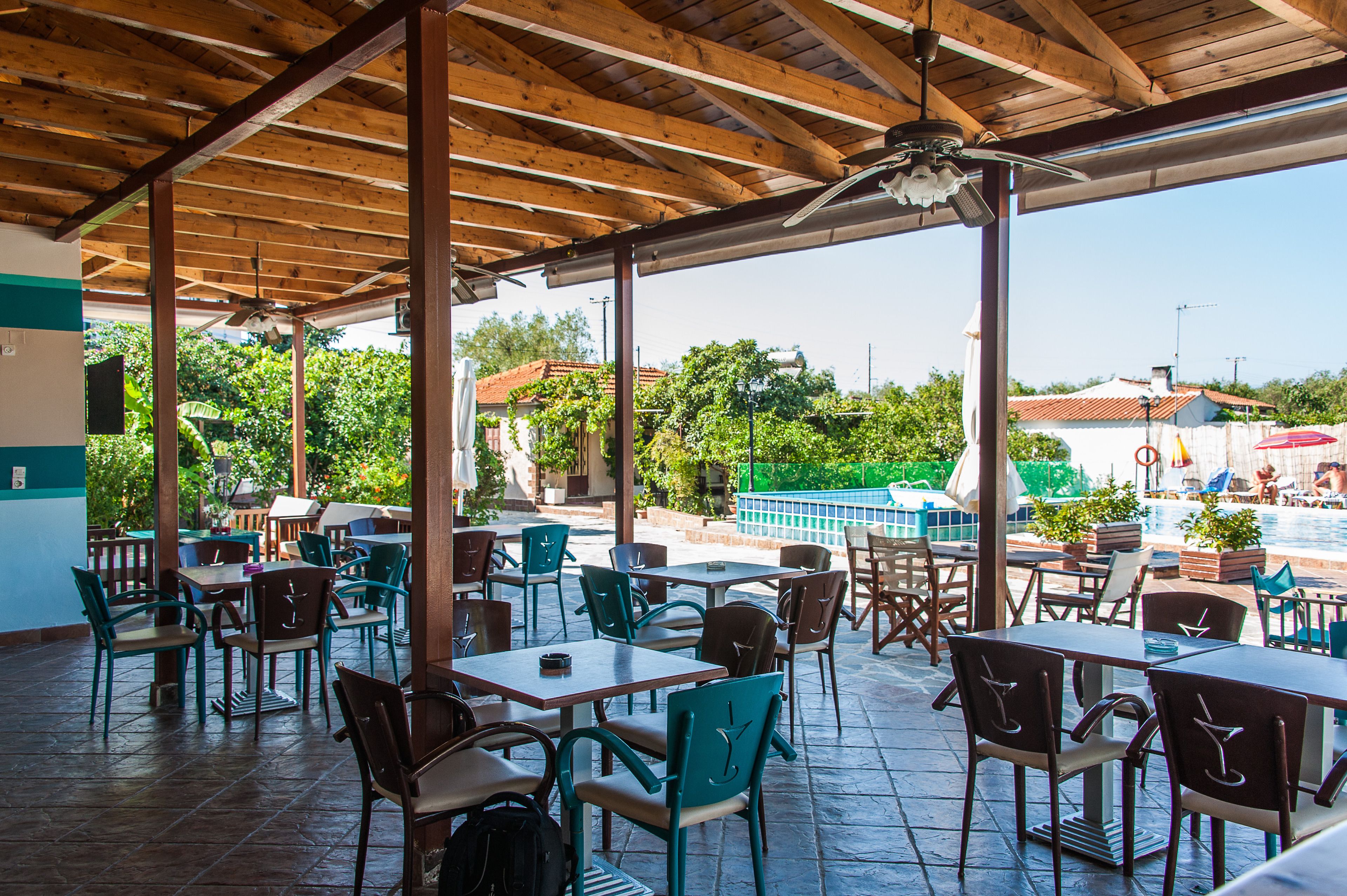 An outdoor patio with wooden tables and chairs, a pool in the background, and a view of surrounding buildings and greenery.