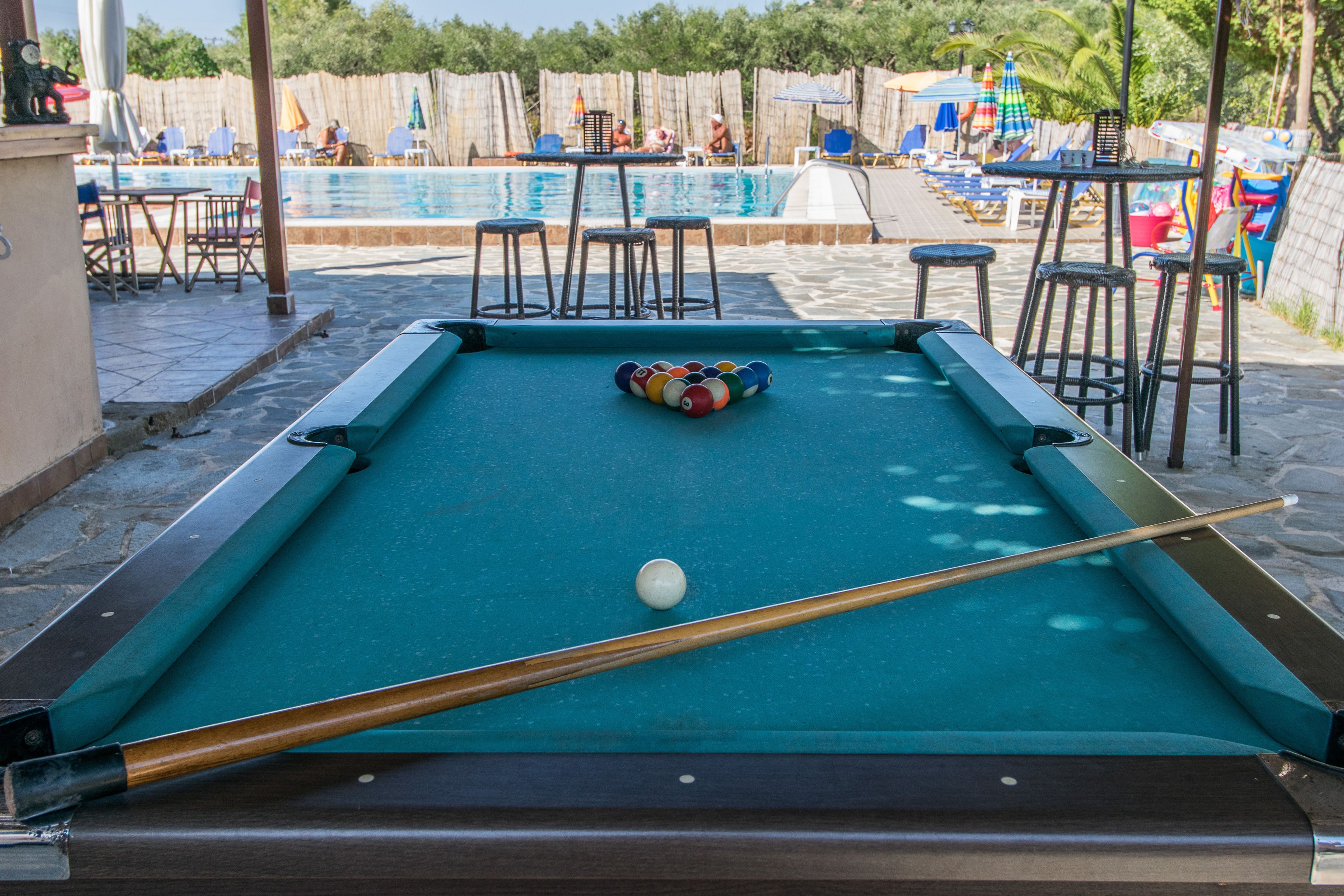 Une table de billard avec une boule et un queue, entourée d’autres tables de billard et de chaises, avec une piscine en arrière-plan.