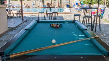 Une table de billard avec une boule et un queue, entourée d’autres tables de billard et de chaises, avec une piscine en arrière-plan.