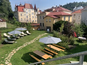 A courtyard with outdoor seating, a stone pathway, and a building with red roofs in the background. - Spa Hotel Devin (Marianske Lazne)