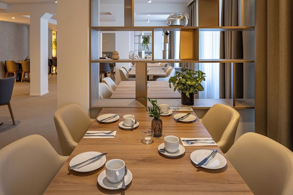 A dining area with a wooden table set for four, surrounded by beige chairs. A vase with a plant is on the table, and there are dining utensils and cups on the tablecloth. - Spa Hotel Devin (Marianske Lazne)