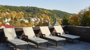 Terraza en la azotea con cuatro sillones de relax y suelo de baldosas, con vistas a una ladera boscosa con edificios.