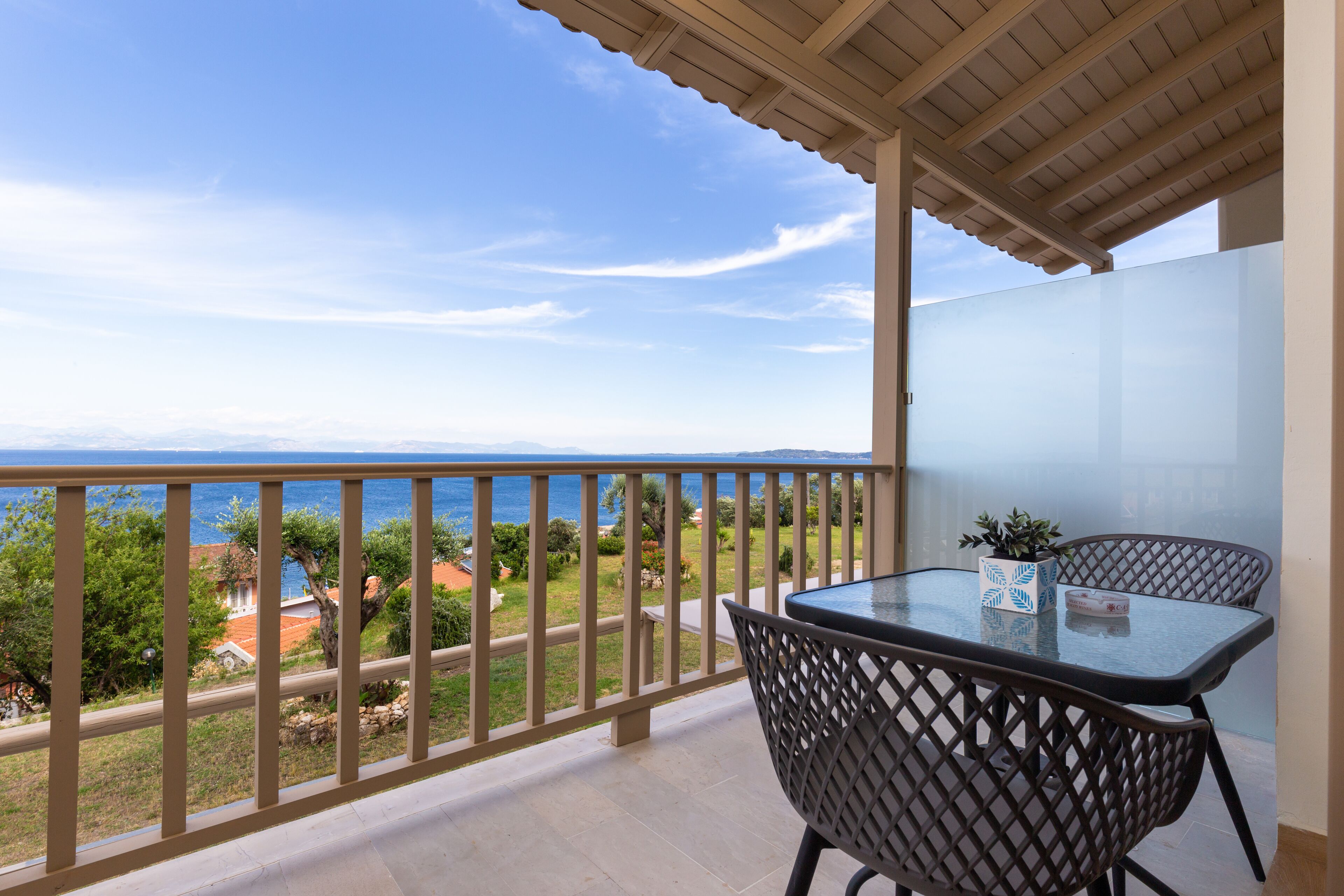 A balcony with a glass table and chairs, overlooking a coastal view with a clear sky.