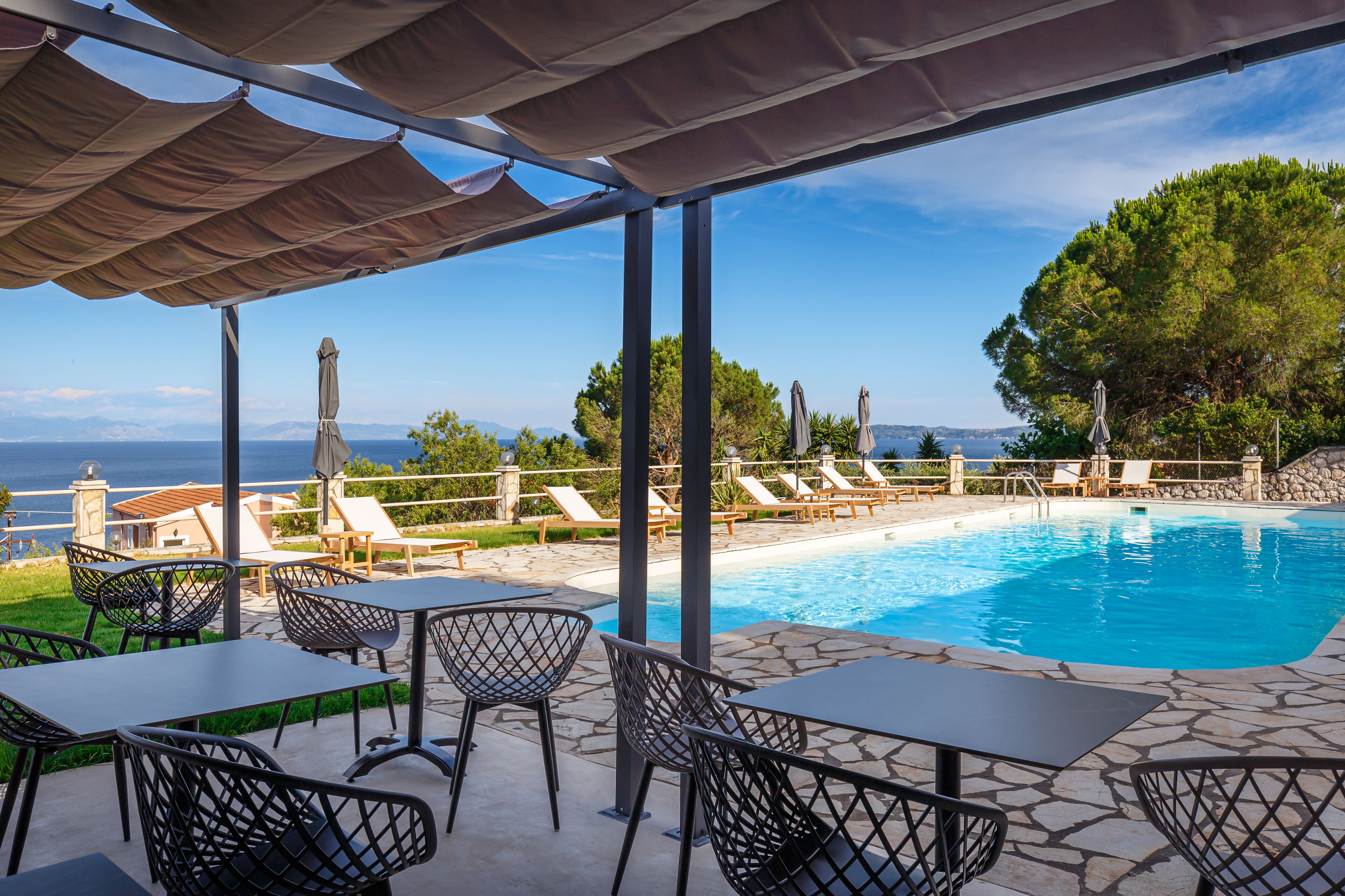 A poolside area with lounge chairs and tables, a clear view of the sea, and a stone pool.
