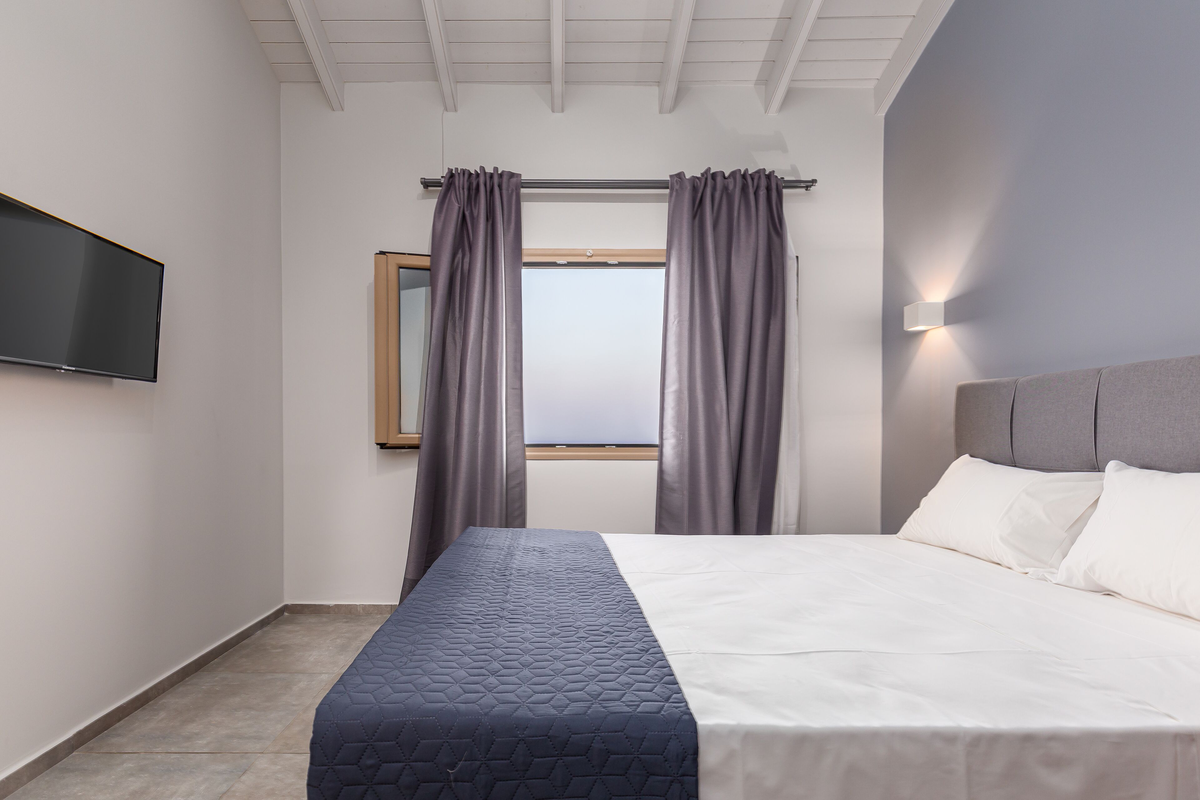 A modern hotel room with a bed, a flat-screen TV, a window with curtains, and a wall-mounted light.