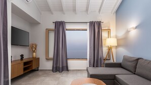 A modern living room with a grey sofa, a wooden coffee table with decorative items, a flat-screen TV, and a view through a window with grey curtains. - Anasa Corfu (Corfu)