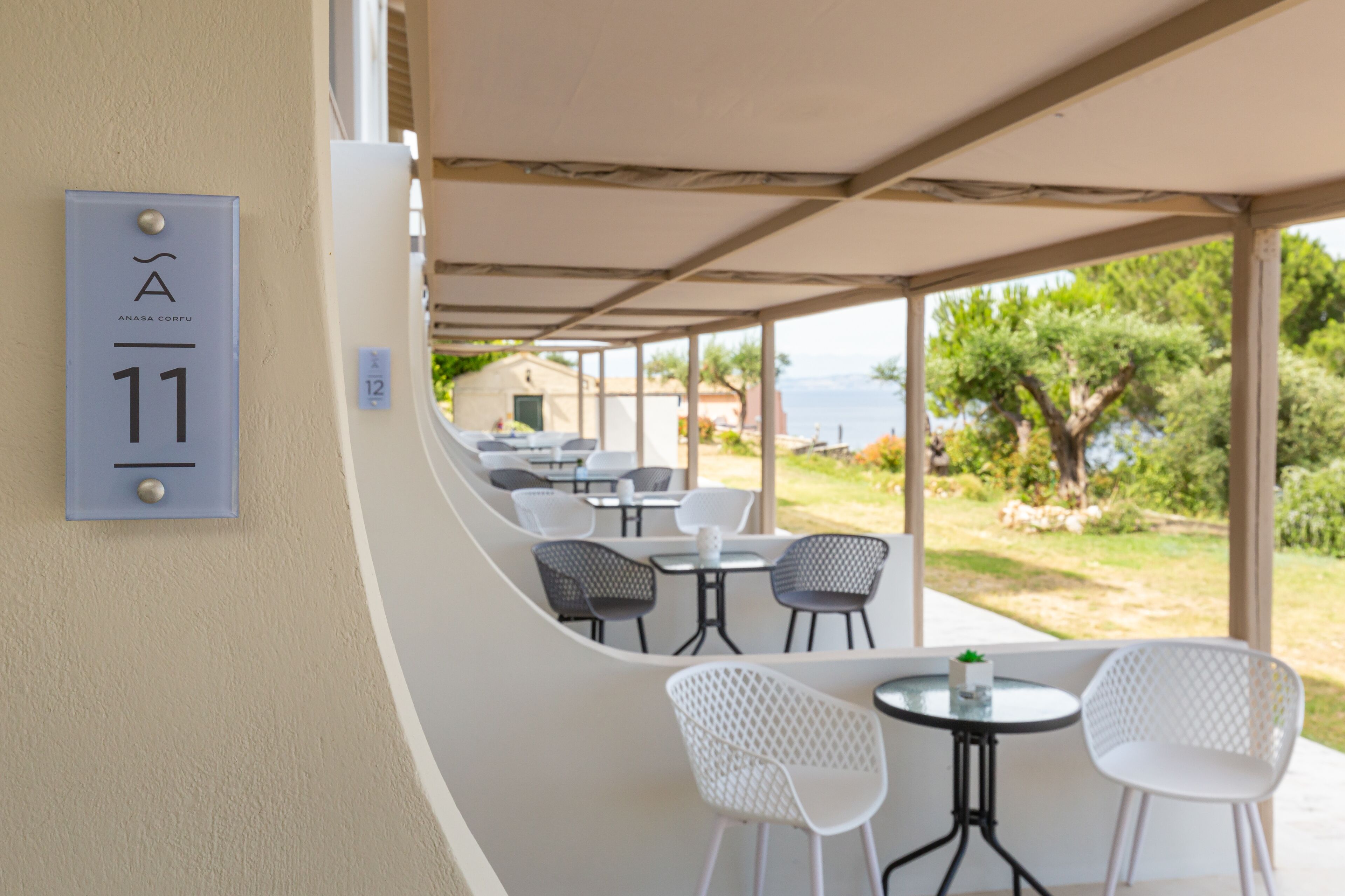 A covered outdoor seating area with tables and chairs, a view of the sea, and a building with a number sign.