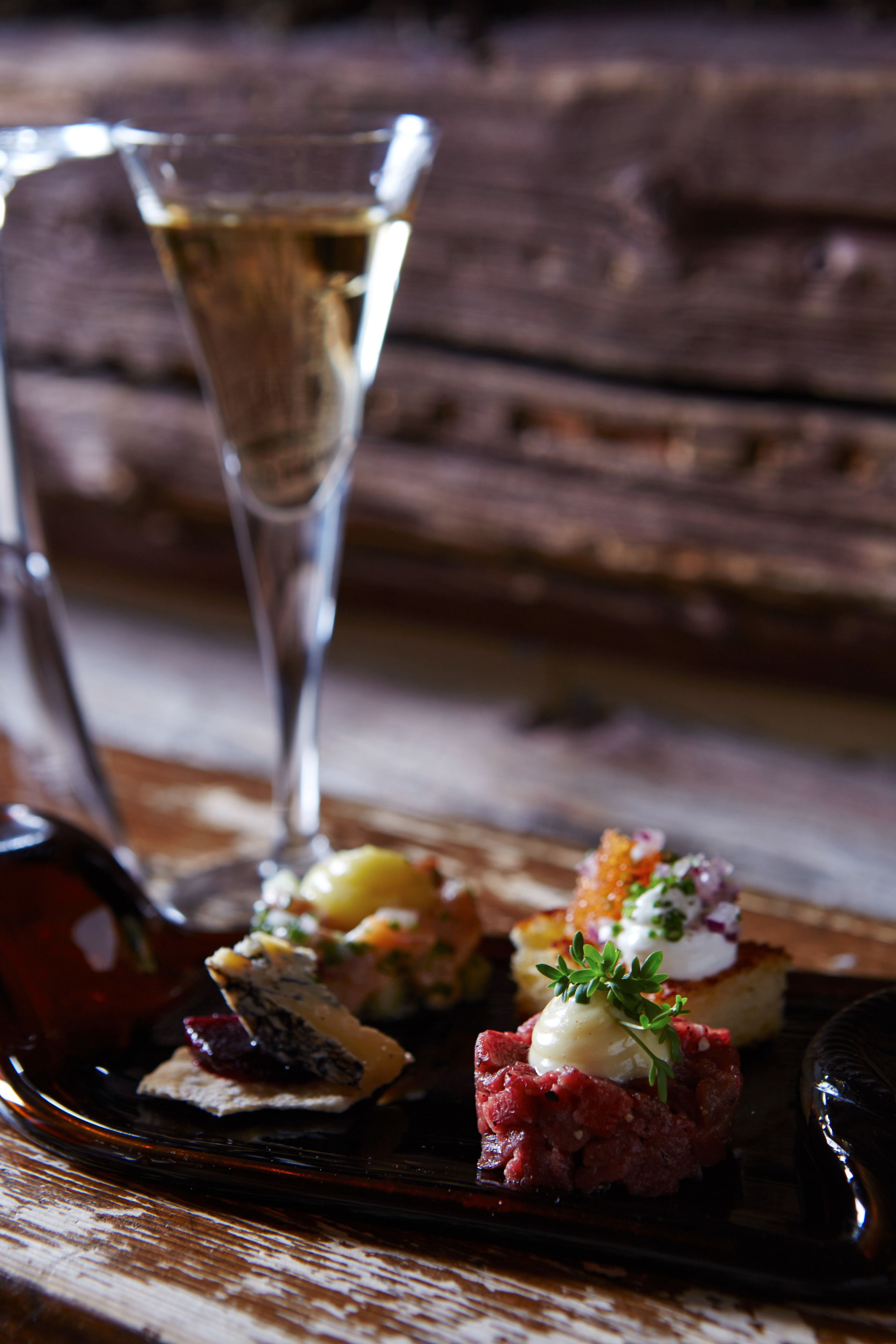 A wooden table with a glass of champagne, a bottle of red wine, and a plate of food.