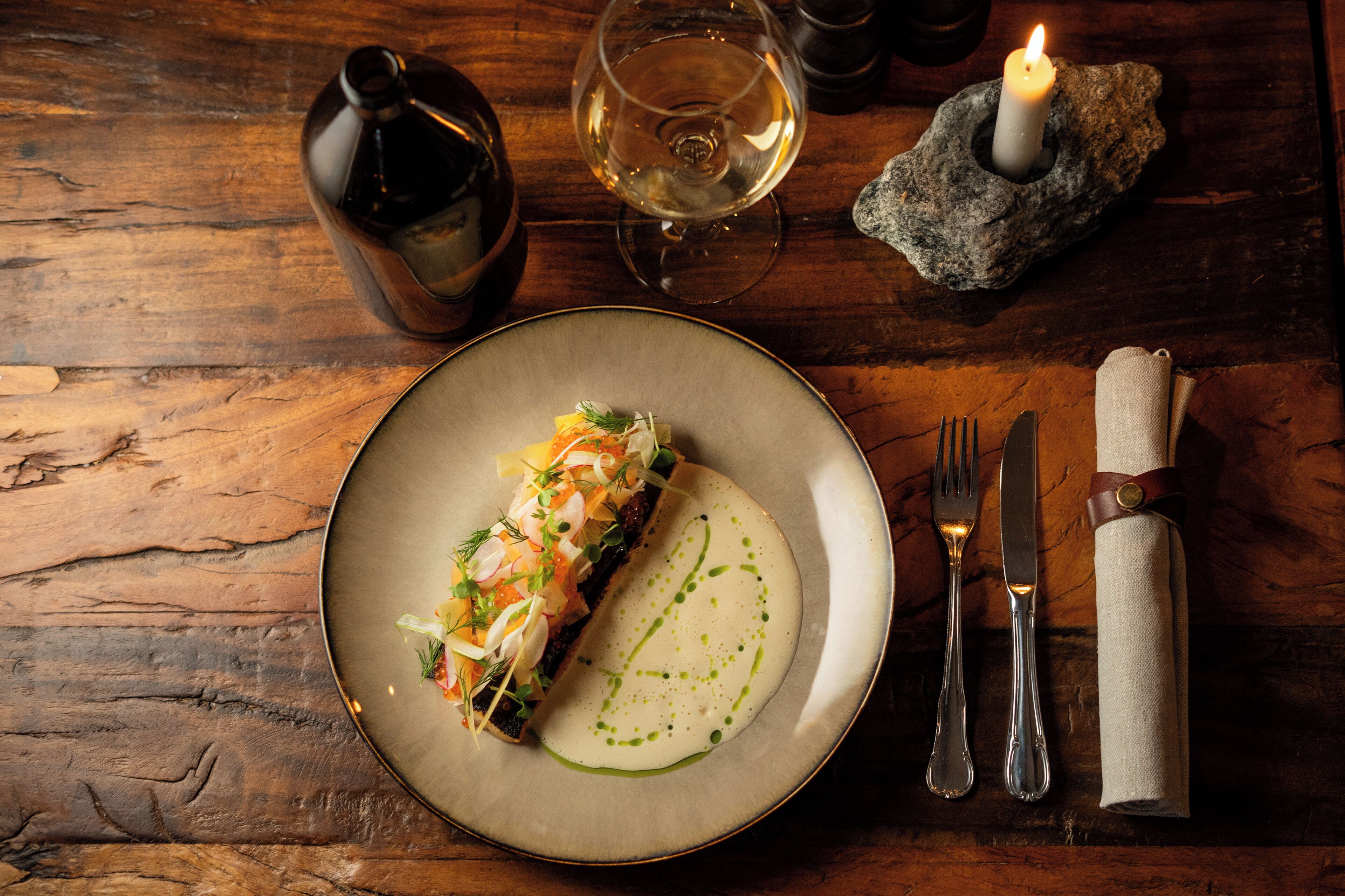 A wooden table with a plate of food, a glass of white wine, a candle, and cutlery.