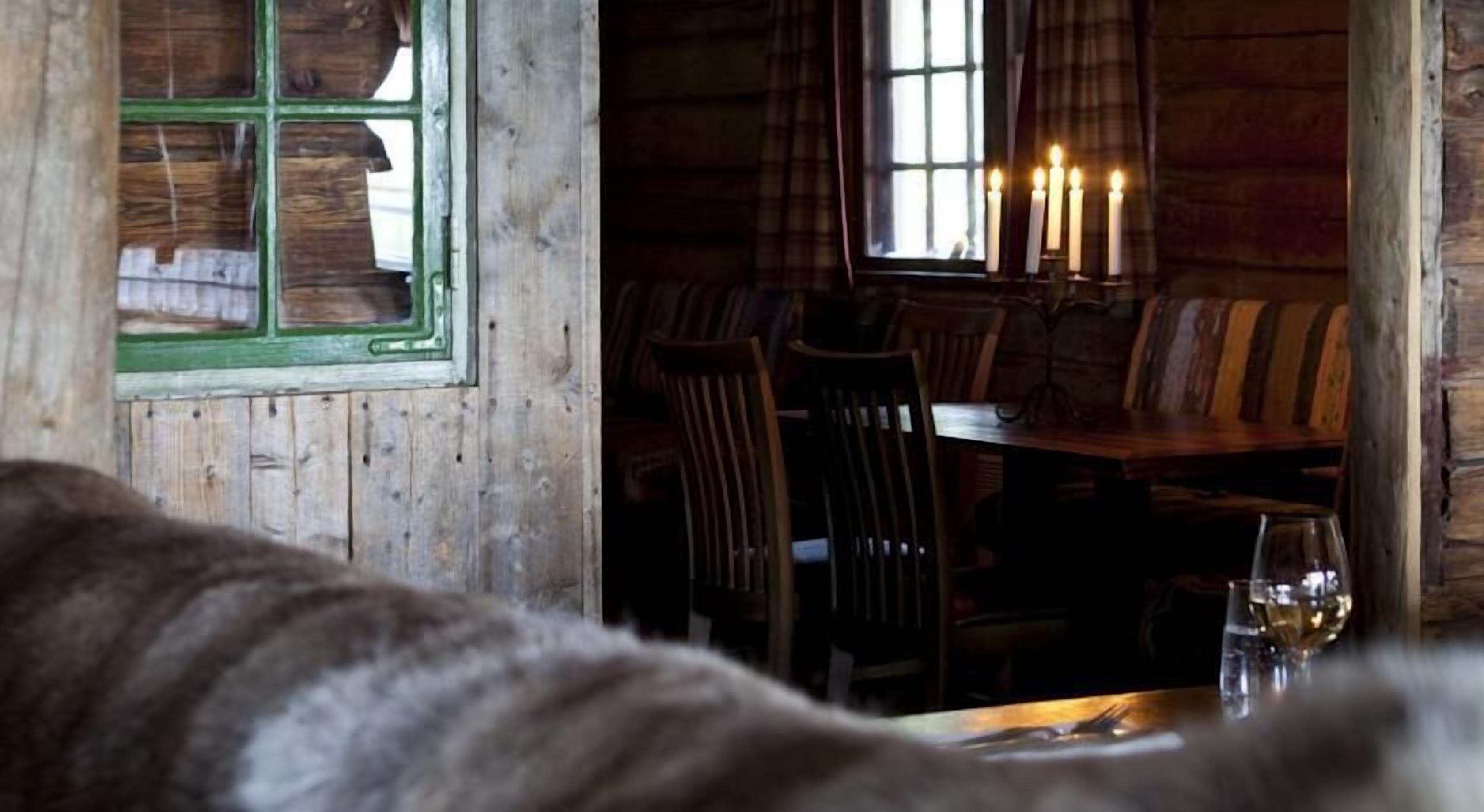 A rustic wooden interior with a dining area featuring a table and chairs, a window with a green frame, and a candlelit ambiance.