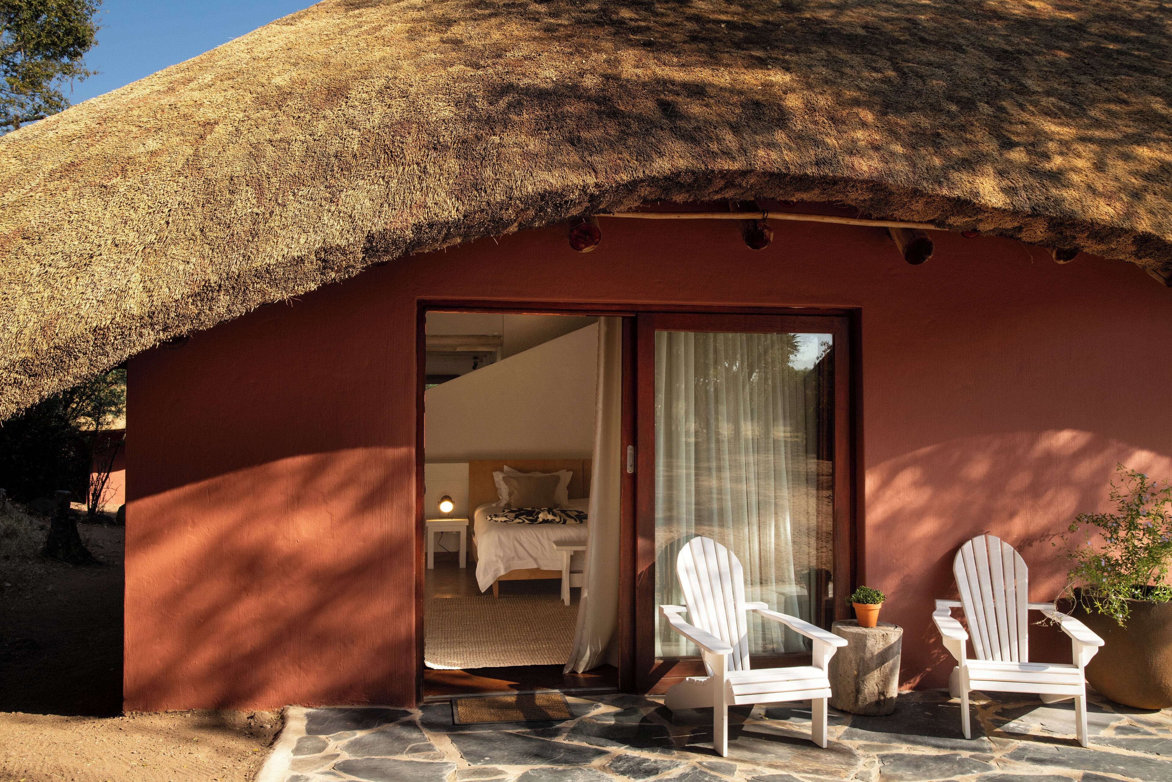 A thatched-roof structure with a bedroom visible inside, two white chairs outside, and a potted plant.