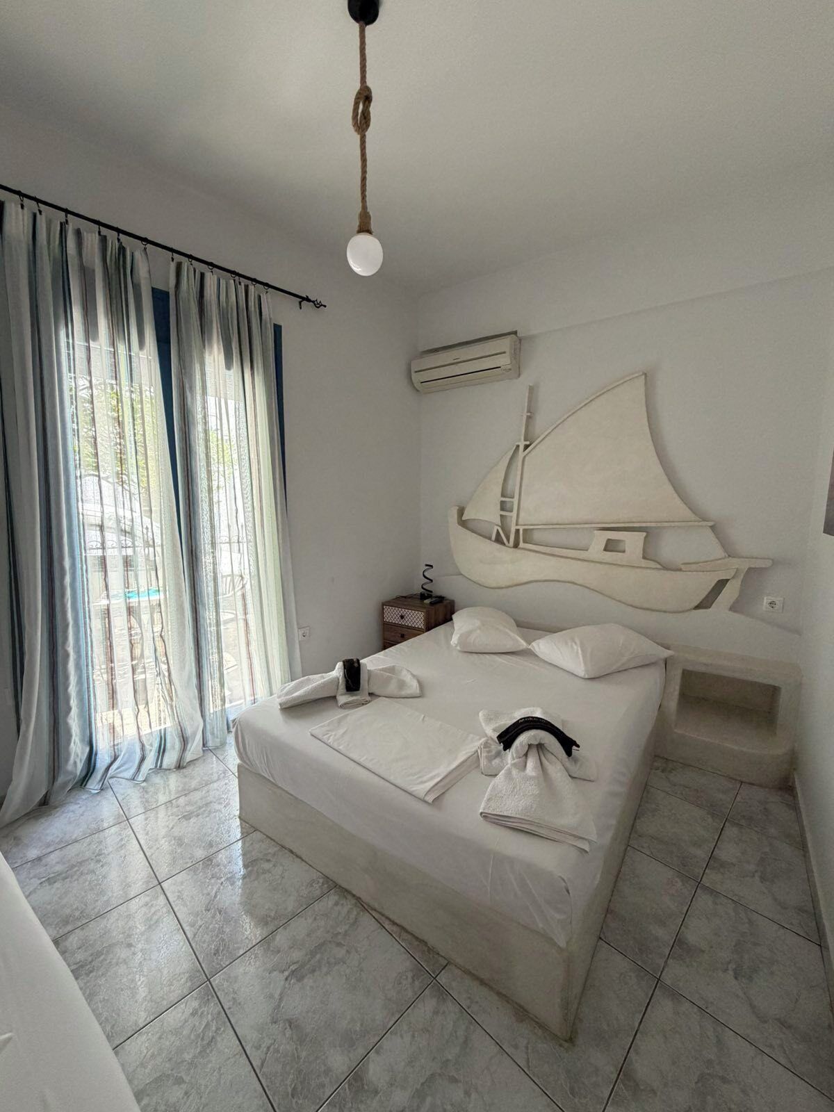 Une chambre d’hôtel avec un lit, une tête de lit ornée d’un motif de voilier, une table de chevet et une fenêtre avec des rideaux.