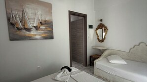 A hotel room with a bed, a sofa, a painting of sailboats, a mirror, and a hanging light fixture. - Foinikas Studios (Kithnos)