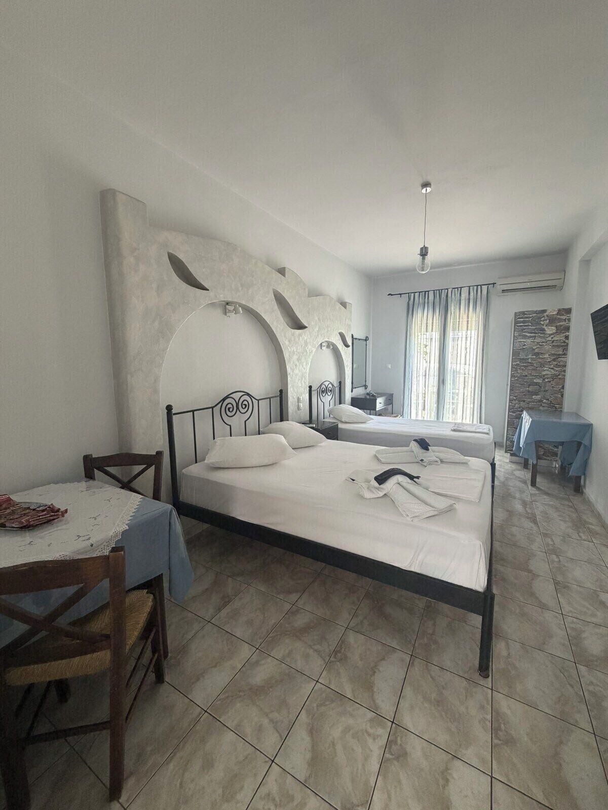 Habitación de hotel con cama, una mesa y sillas, una mesita auxiliar y un ventanal con cortinas.