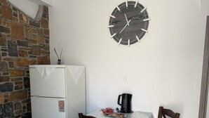 A small dining area with a table, two chairs, a clock on the wall, a refrigerator, and a stone wall. - Foinikas Studios (Kithnos)