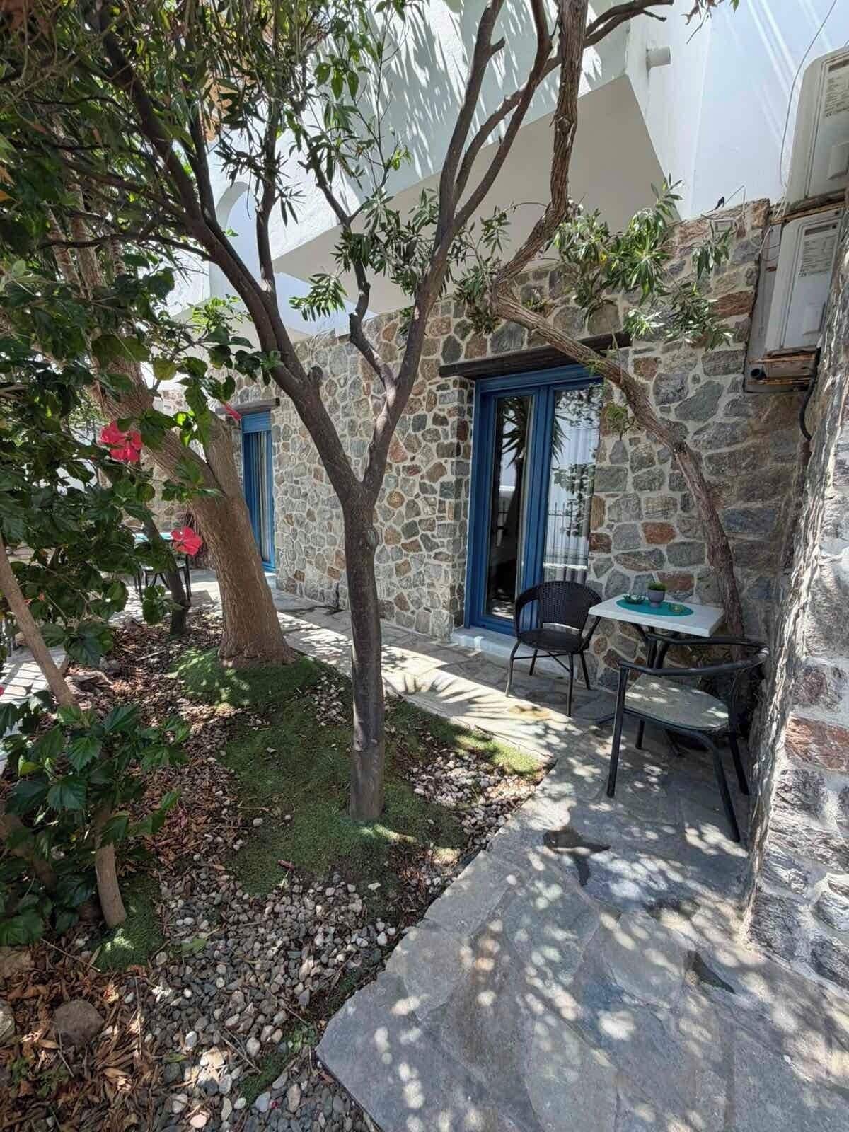 Un edificio de piedra con una puerta azul, un pequeño espacio al aire libre con mesa y sillas, y un árbol de hojas verdes.