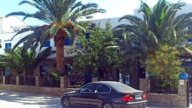 A black car parked in front of a building with stone and stucco walls, palm trees, and a blue door. - Foinikas Studios (Kithnos)