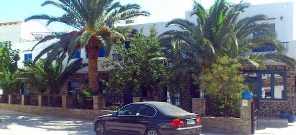 A black car parked in front of a building with stone and stucco walls, palm trees, and a blue door. - Foinikas Studios (Kithnos)