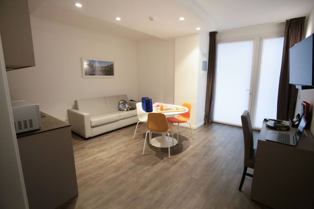 A modern living room with a sofa, a dining table with chairs, a flat-screen TV, and a laptop on a desk.