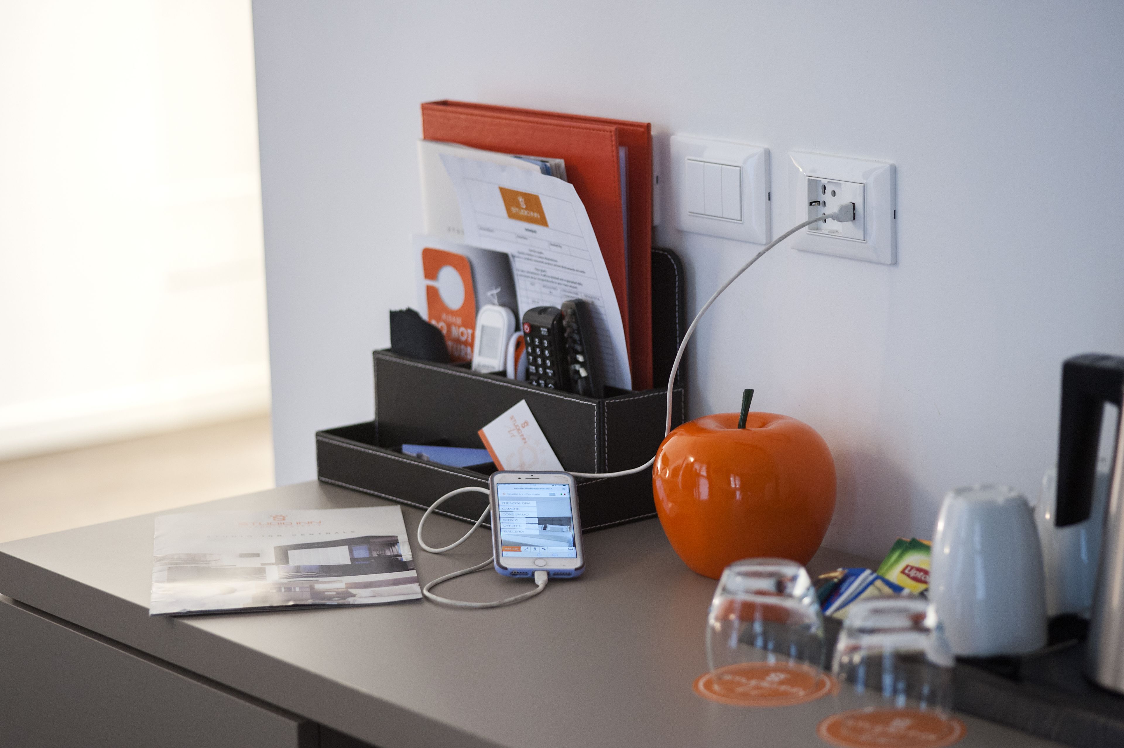 A desk with a smartphone charging, a red folder, a black organizer, and a glass of orange juice.