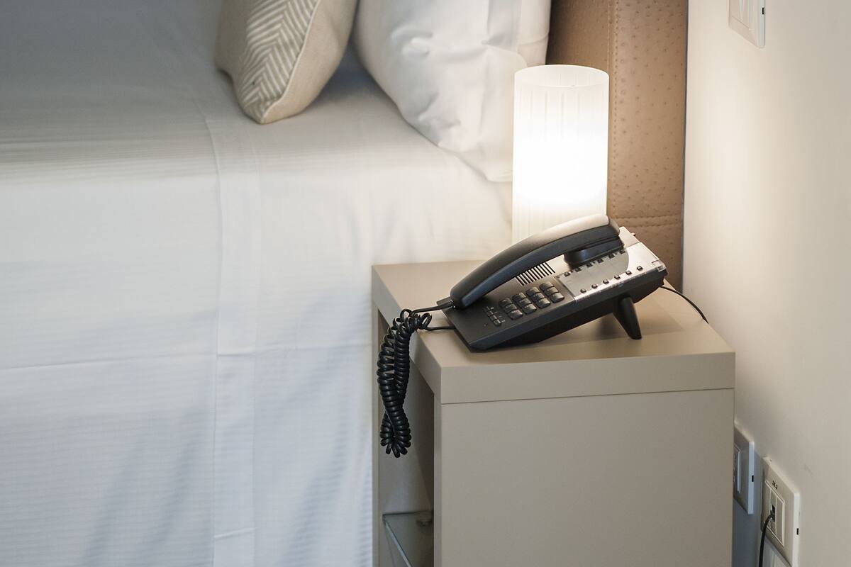 A neatly made bed with white linens and several pillows, a bedside lamp, and a telephone on a small table beside the bed.