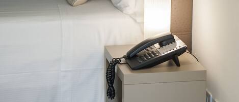 A neatly made bed with white linens and several pillows, a bedside lamp, and a telephone on a small table beside the bed.