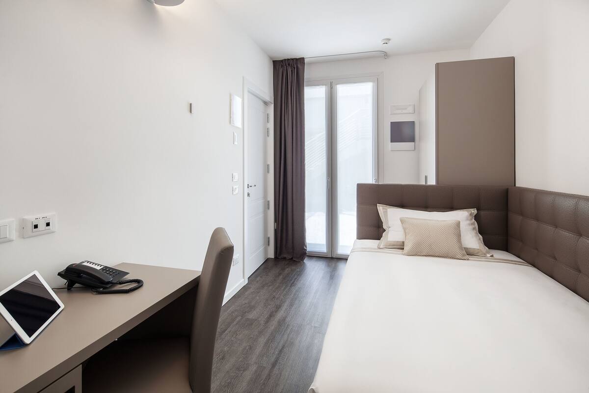 A hotel room with a bed, a desk with a phone and tablet, a chair, and a view of the outside through a door.