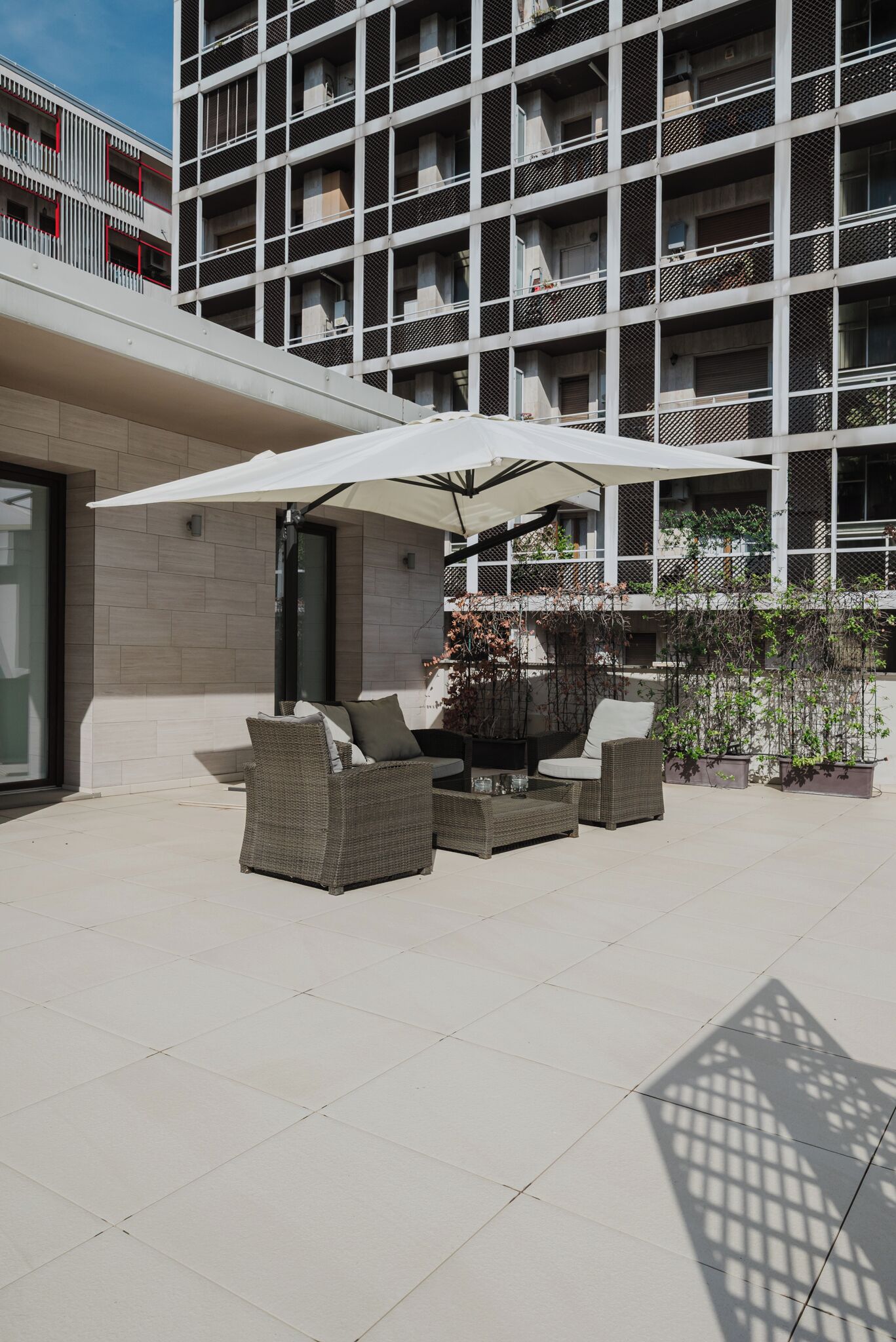 A modern outdoor seating area with wicker furniture and a large umbrella, adjacent to a multi-story building with balconies.