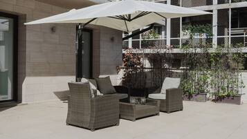 A modern outdoor seating area with wicker furniture and a large umbrella, adjacent to a multi-story building with balconies.
