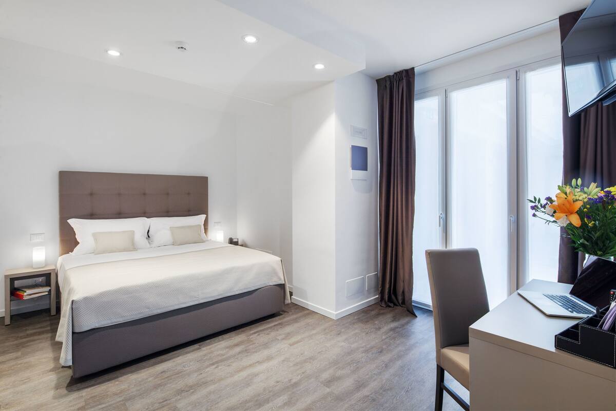 A modern hotel room with a large bed, a desk with a laptop, a TV, and a window with curtains.