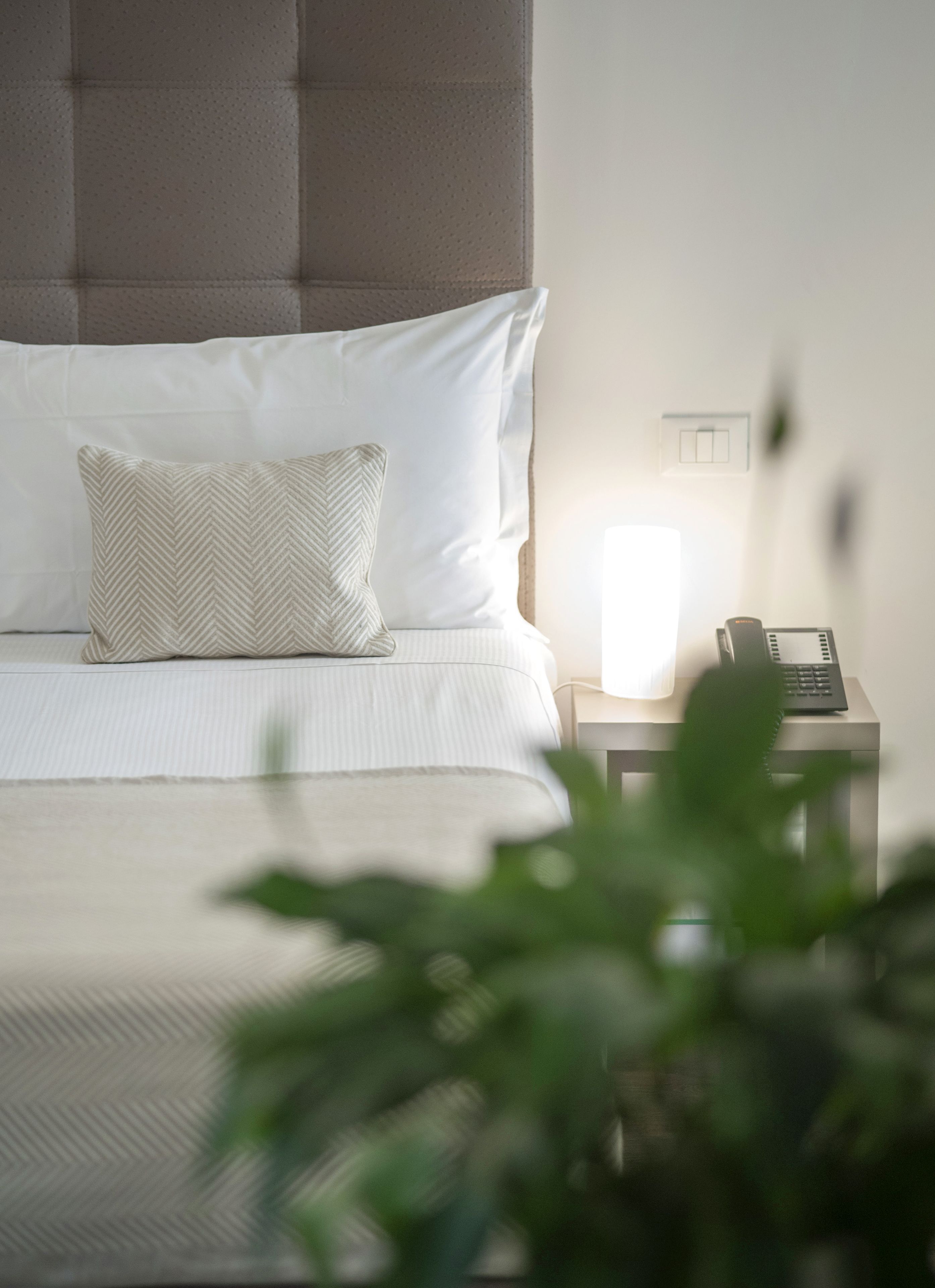 Ein ordentlich gemachtes Bett mit weißen Leinen und einem Kissen in Fischgrätmuster, ein Nachttisch mit Telefon und ein Lichtschalter an der Wand.