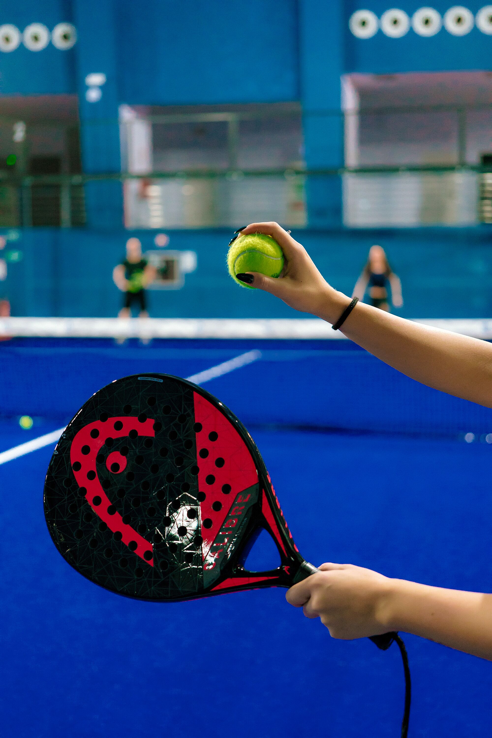 Een hand die een tennisracket vasthoudt, met een rood en zwart ontwerp, een tennisbal en een blauwe tennisbaan op de achtergrond.