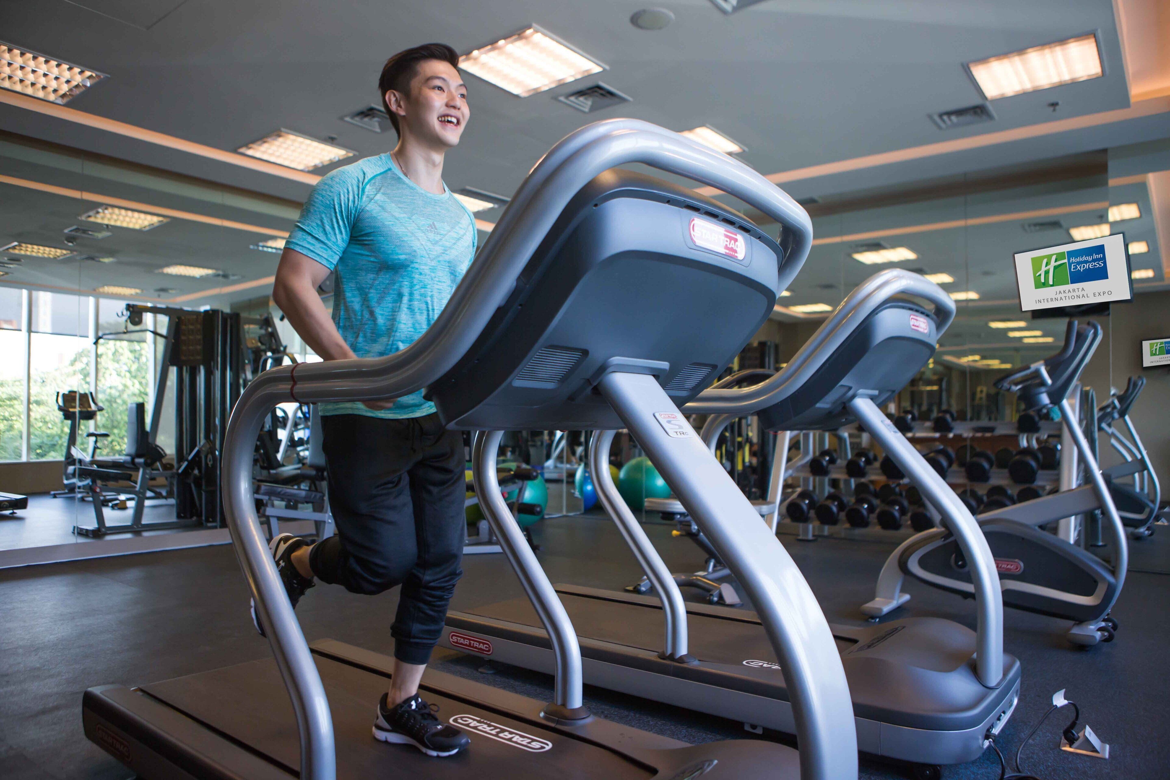 A person running on a treadmill in a gym with exercise equipment and a sign for Holiday Inn Express Jakarta International Expo.